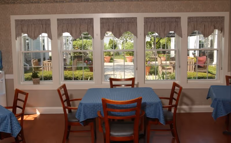 A dining area with wooden chairs and tables covered with blue tablecloths. Large windows with checkered valances provide a view of an outdoor garden area with potted plants and a person sitting on a bench.