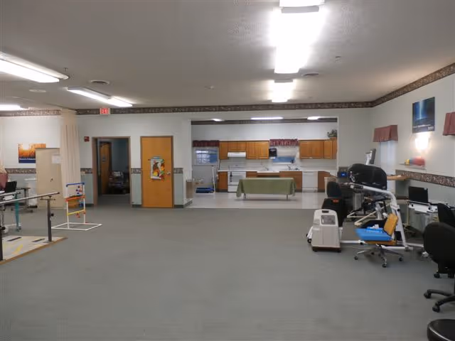 Large open activity/therapy room with a kitchenette at the back, exercise/therapy equipment and chairs along the sides.