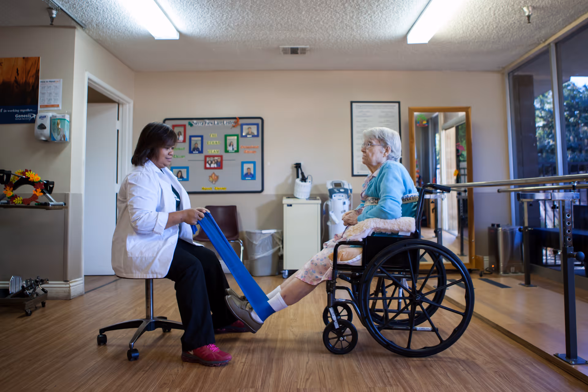 A healthcare professional assists an elderly woman in a wheelchair with a leg exercise using a blue resistance band in a therapy room with wooden floors and large windows.