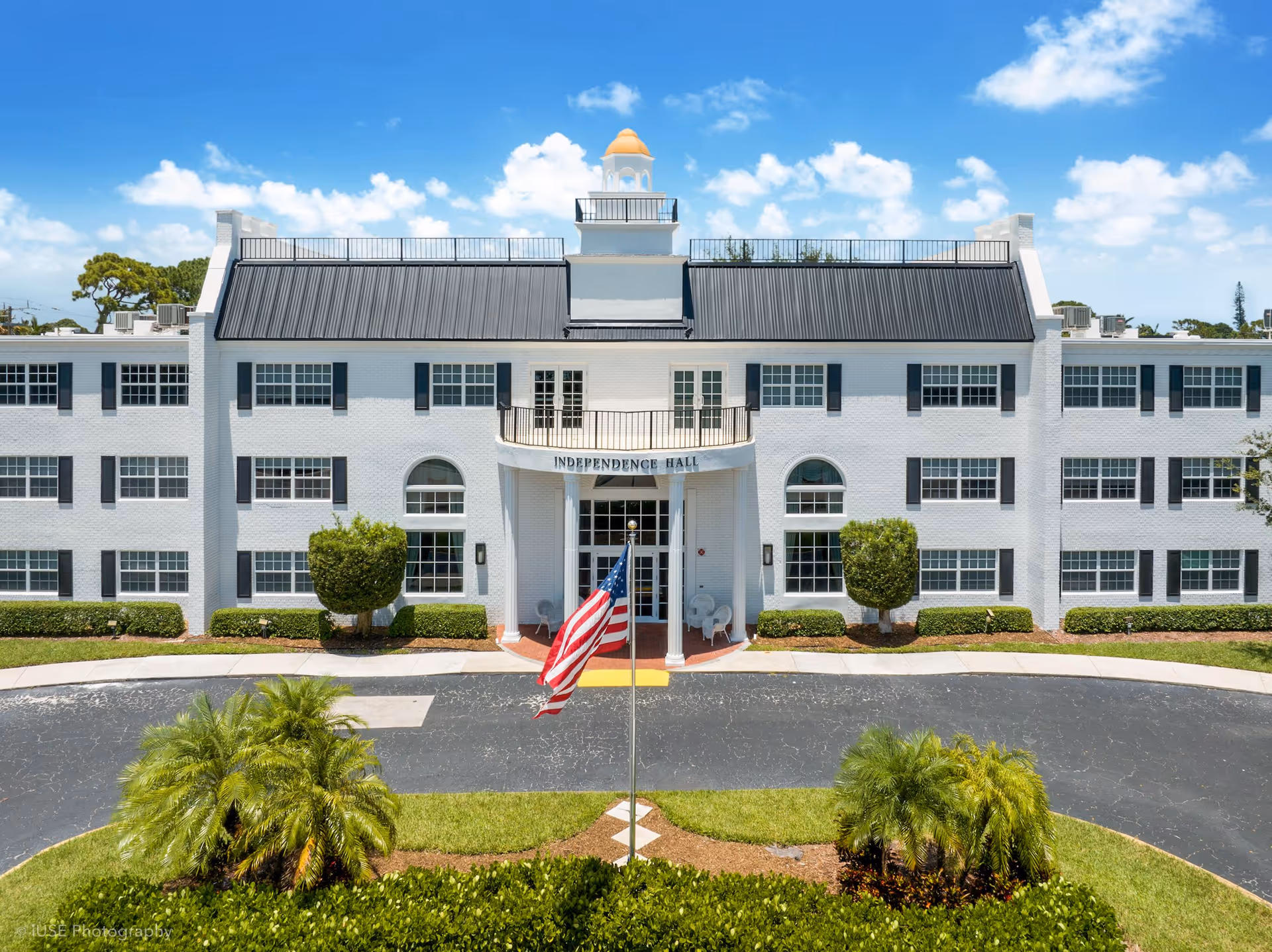 Front exterior view of Independence Hall Senior Living building, a large white three-story structure with black shutters and a black roof. There is a balcony above the entrance with the name Independence Hall displayed. An American flag is flying on a flagpole in front of the building, surrounded by green landscaping and palm trees under a blue sky with scattered clouds.