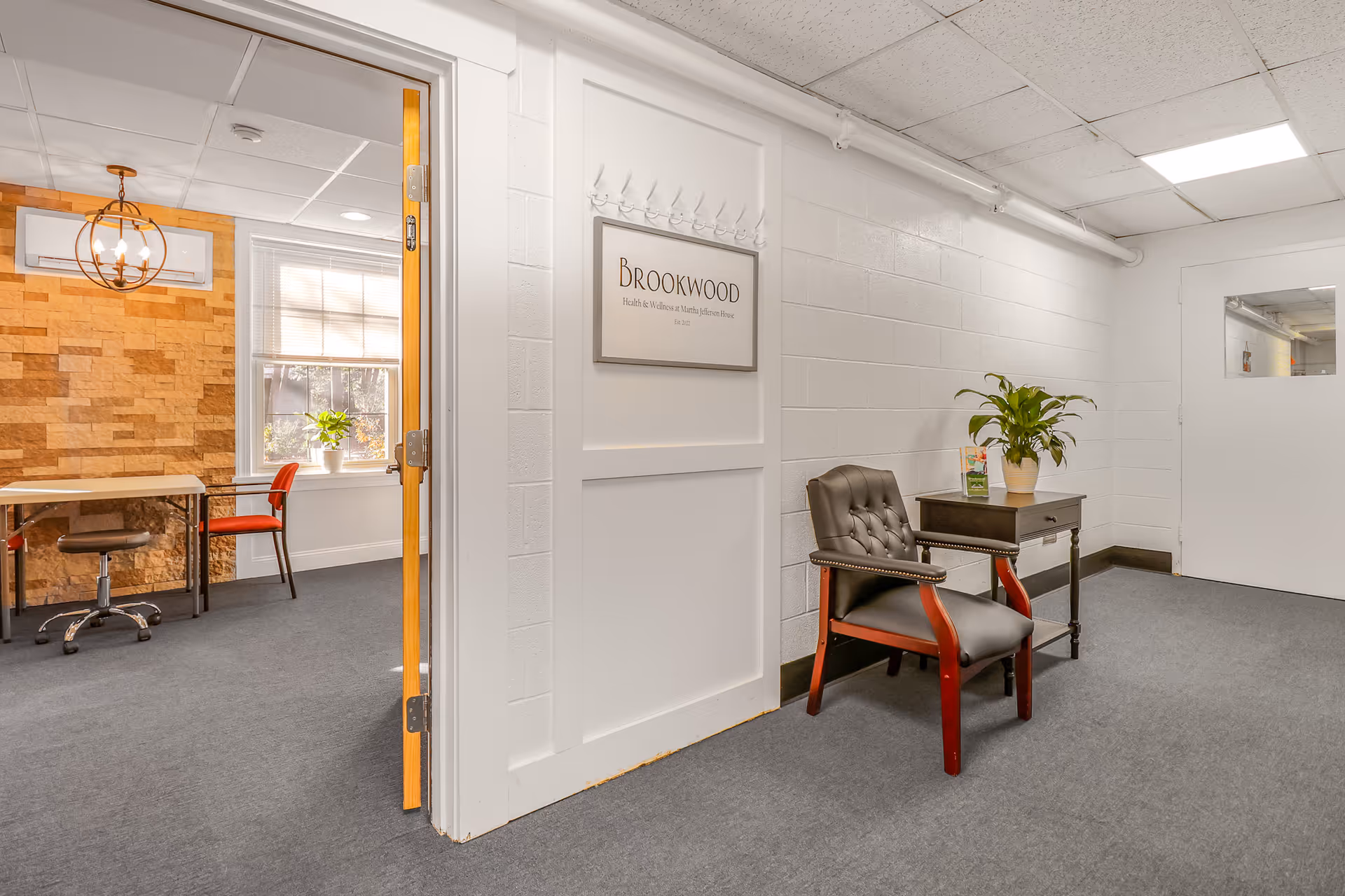 A hallway with gray carpet and white walls leading to a room with a wooden accent wall and a window with blinds. In the hallway, there is a black cushioned chair with wooden armrests next to a small dark wooden table holding a potted plant and a brochure. A sign on the wall reads 'Brookwood Health & Wellness at Martha Jefferson House'.