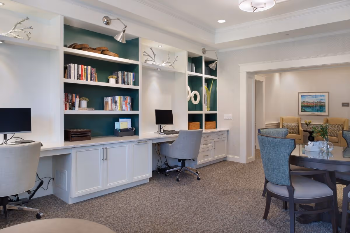 A bright communal lounge with built-in desks and bookshelves, computer workstations, a round table with chairs, and a seating area in the background.