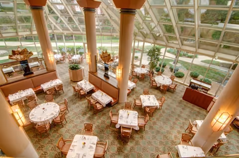 A spacious dining room with multiple round and square tables covered with white tablecloths and set with dining ware. The room features large floor-to-ceiling windows with a view of greenery outside, tall columns, and warm lighting fixtures on the walls. There are plants and decorative elements placed around the room.