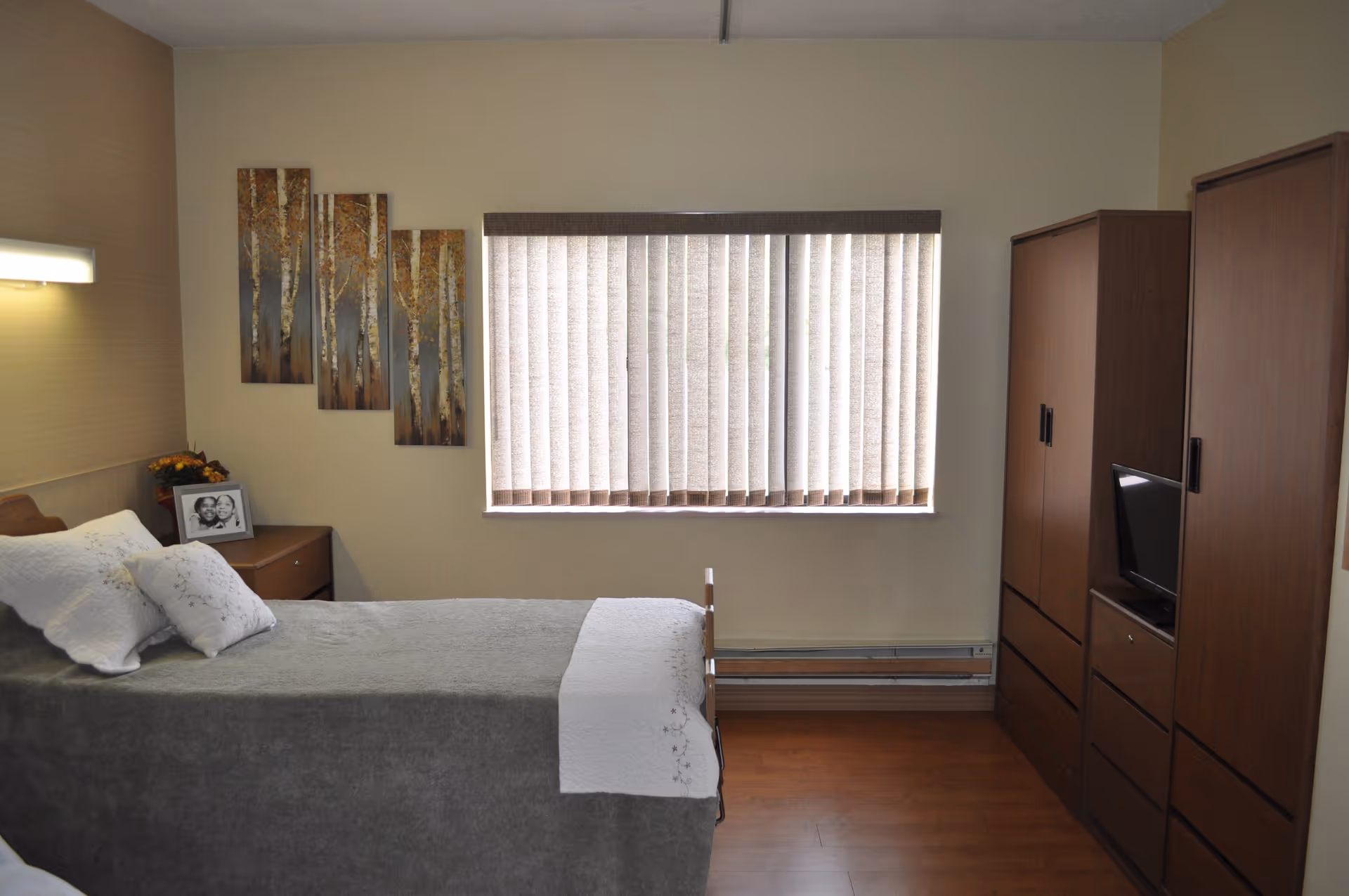 A neatly furnished bedroom with a single bed, bedside table, vertical-blind window and wooden wardrobes.