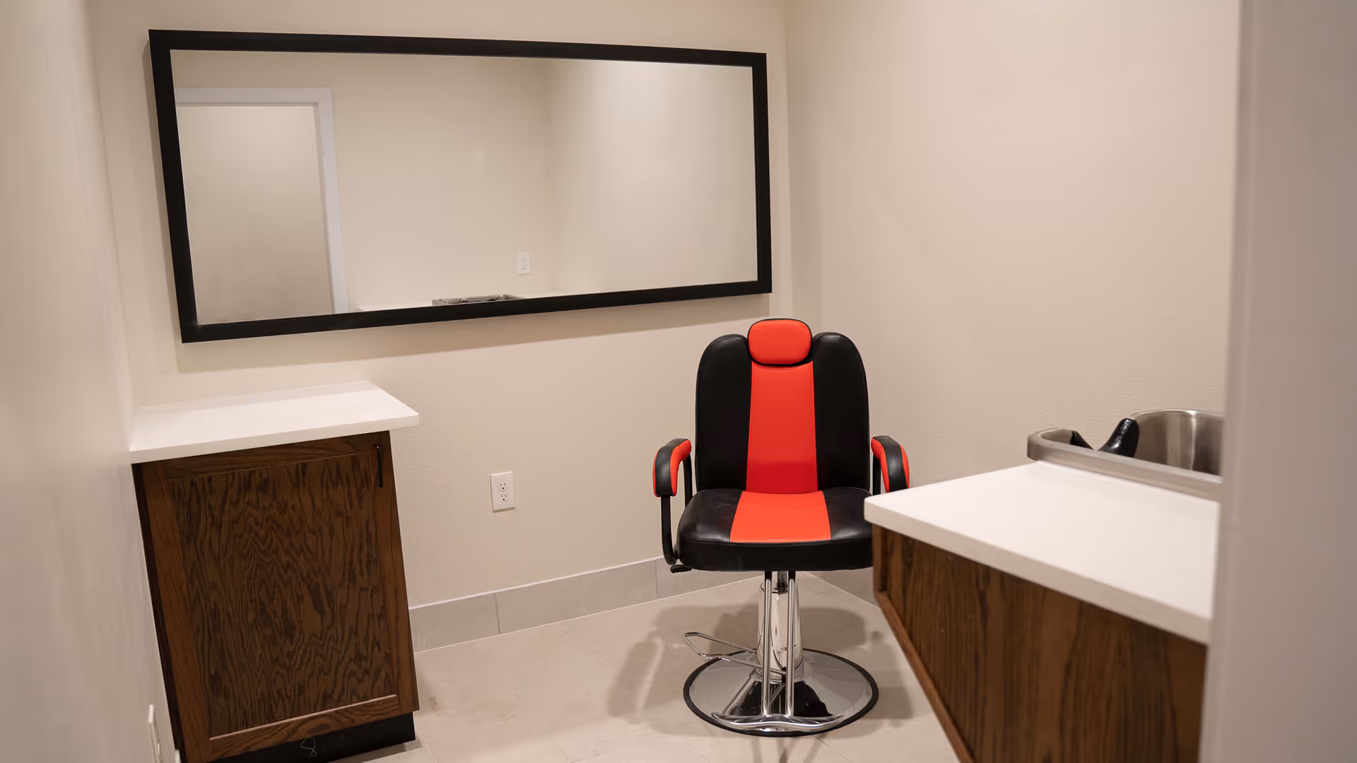 A small room with a black and red salon chair in front of a large rectangular mirror mounted on the wall. There are wooden cabinets with white countertops on either side of the chair, and a small stainless steel sink is visible on the right countertop. The walls are light-colored and the floor is tiled.