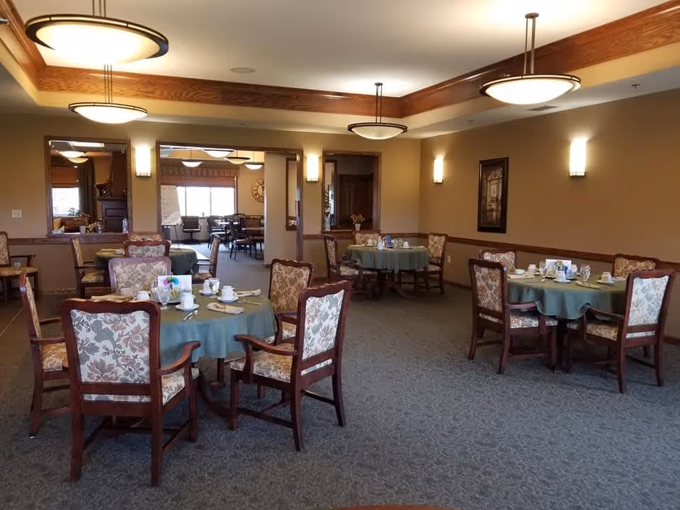 A dining room in an assisted living facility with several round tables covered with green tablecloths. Each table is set with cups, plates, napkins, and silverware. The chairs have wooden frames and floral upholstered backs and seats. The room has beige walls, carpeted floors, and ceiling lights with wooden trim accents. There are mirrors on one wall and a framed picture on another.