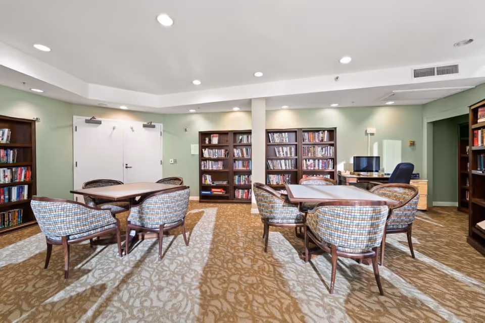 A well-lit common room with tables and upholstered chairs, bookshelves lining the walls, and a desk with a computer.
