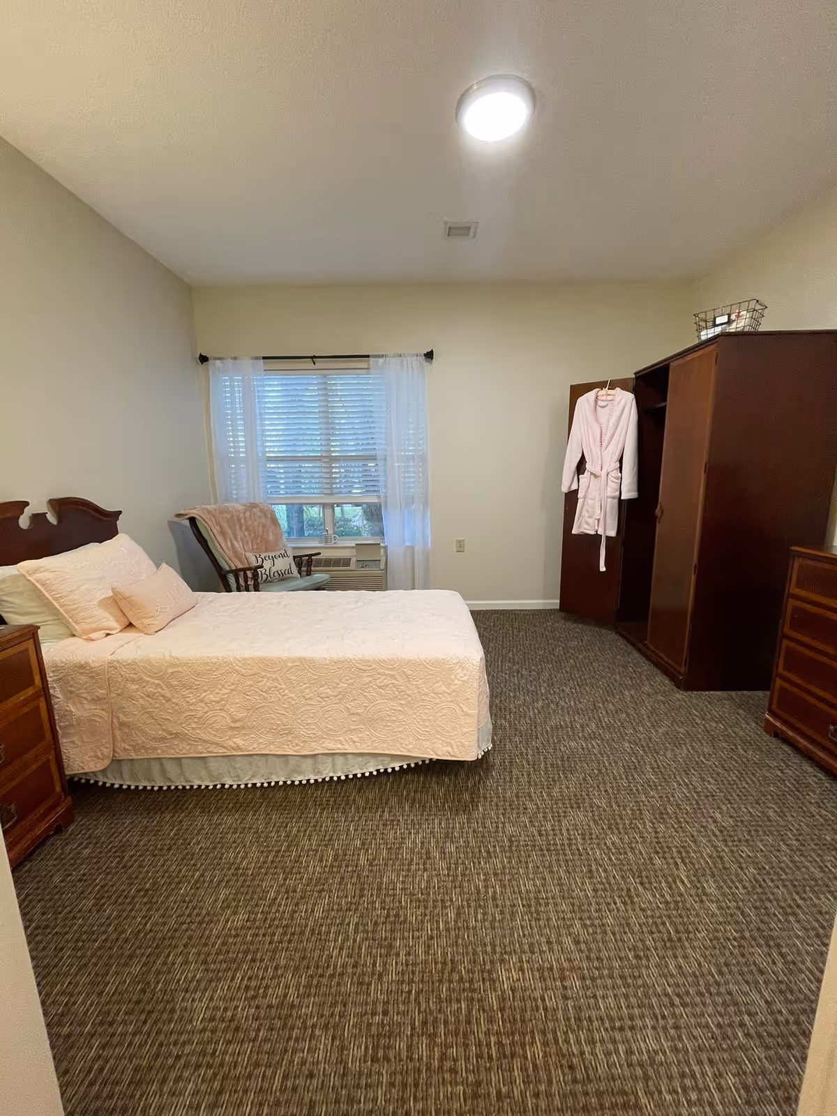 Bright assisted-living bedroom with a neatly made twin bed, wooden dressers and wardrobe, a robe hanging on the wardrobe, and a window with sheer curtains.