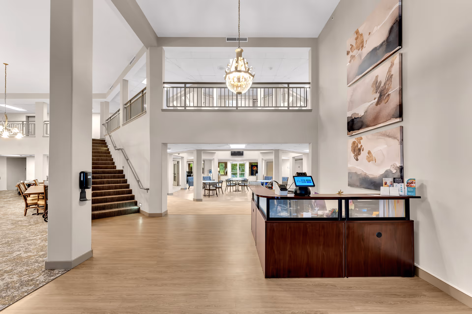 Interior view of a senior living facility lobby area with a wooden reception desk on the right, a staircase with carpeted steps on the left, and a spacious common area with tables and chairs in the background. The walls are painted light beige, and there are three abstract paintings hanging vertically on the right wall. A chandelier hangs from the ceiling in the center.