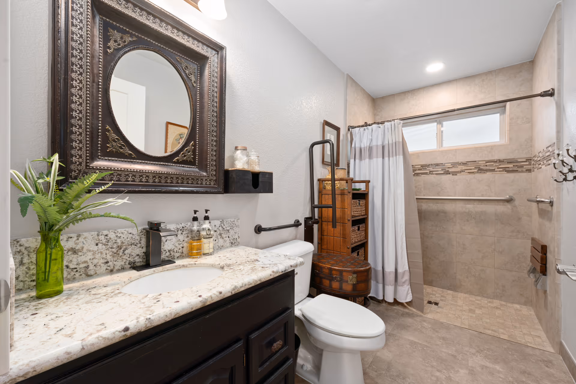 A well-lit bathroom featuring a granite countertop with a sink, a decorative framed mirror above, and a green vase with plants. There is a toilet next to the counter, a wooden storage unit with baskets, and a walk-in shower with a curtain and grab bars. The walls and floor are tiled in neutral tones.