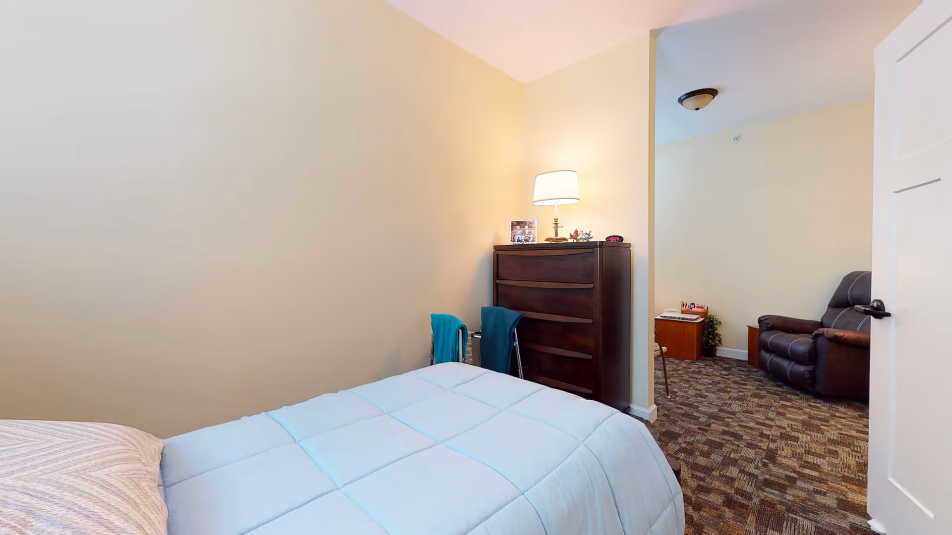 A cozy bedroom with a bed covered in a light blue comforter and a pillow. Next to the bed is a dark wooden dresser with a lamp on top, along with some small decorative items. The room has beige walls and carpeted flooring. Through an open door, a small sitting area is visible with a brown recliner chair, a small table, and some plants.