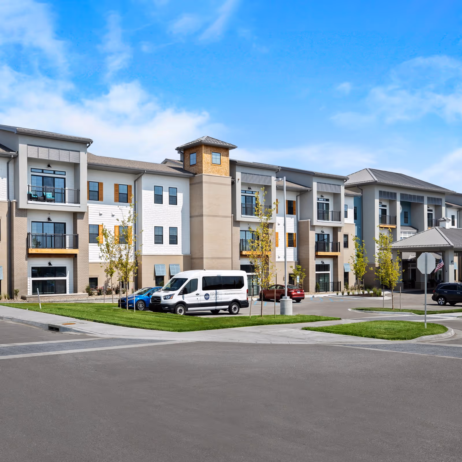 Exterior view of a modern three-story senior living facility building with balconies, large windows, and a covered entrance. Several cars and a white van with the Novel Place logo are parked in the parking lot. The sky is clear and blue with some clouds.