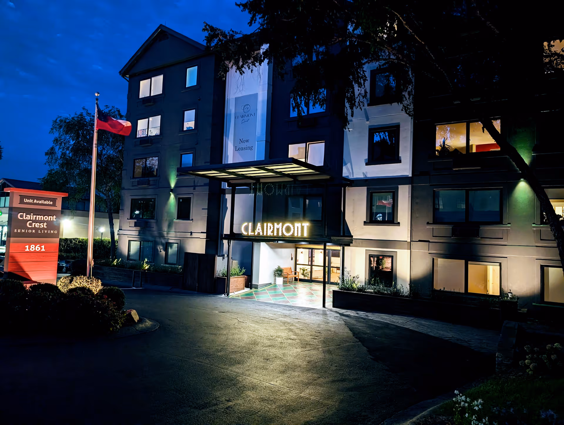 Night view of Clairmont Crest senior living facility building entrance with illuminated sign and a flagpole with a flag. The building has multiple windows lit from inside and a sign near the driveway entrance that reads 'Unit Available Clairmont Crest Senior Living 1861'.