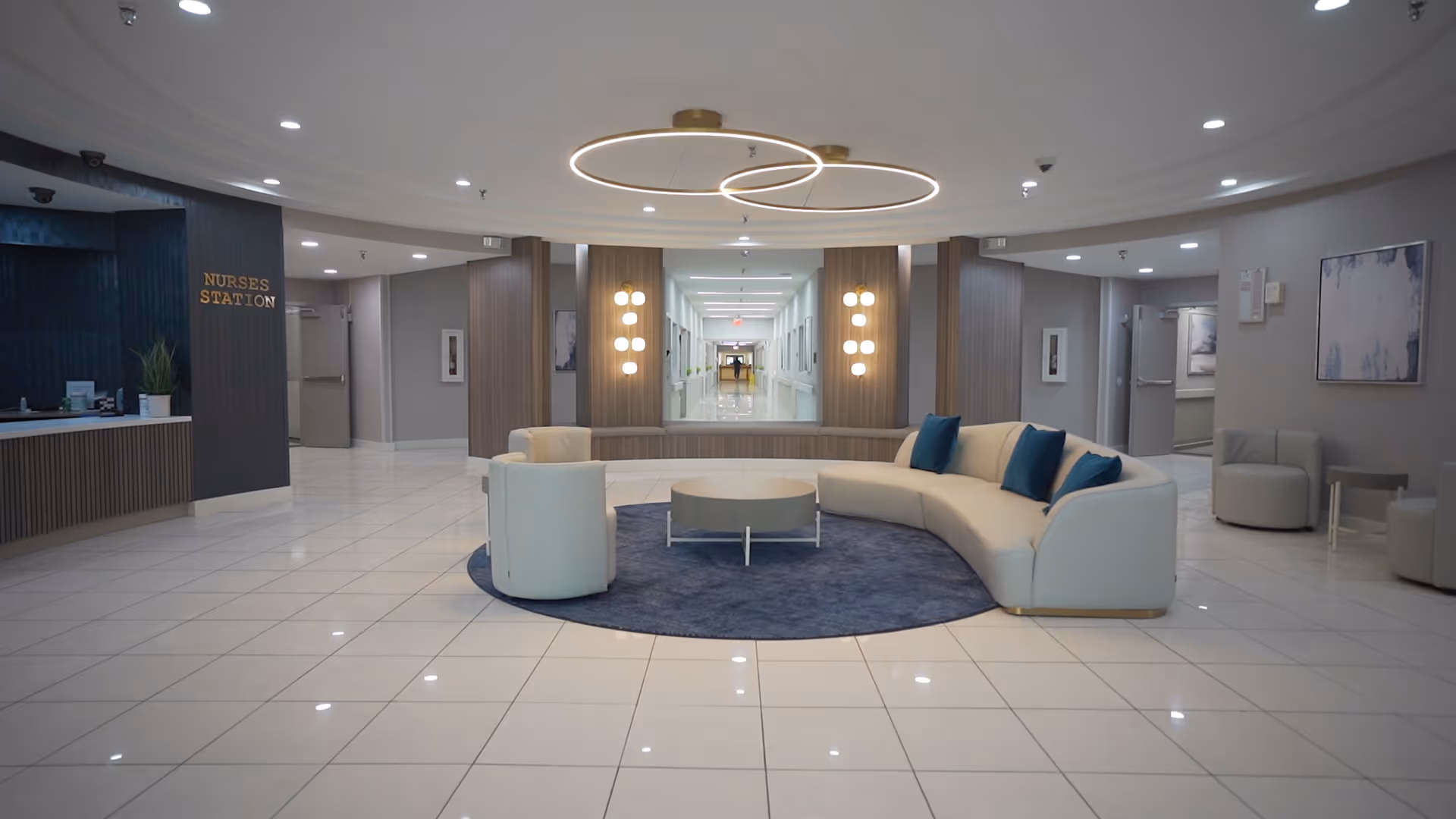 Bright modern lobby with a curved white sofa on a round blue rug, a nurses station to the left, and a long hallway beyond.