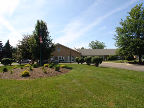 Low single-story senior living building with a landscaped lawn, flagpole, driveway, and trees under a blue sky.