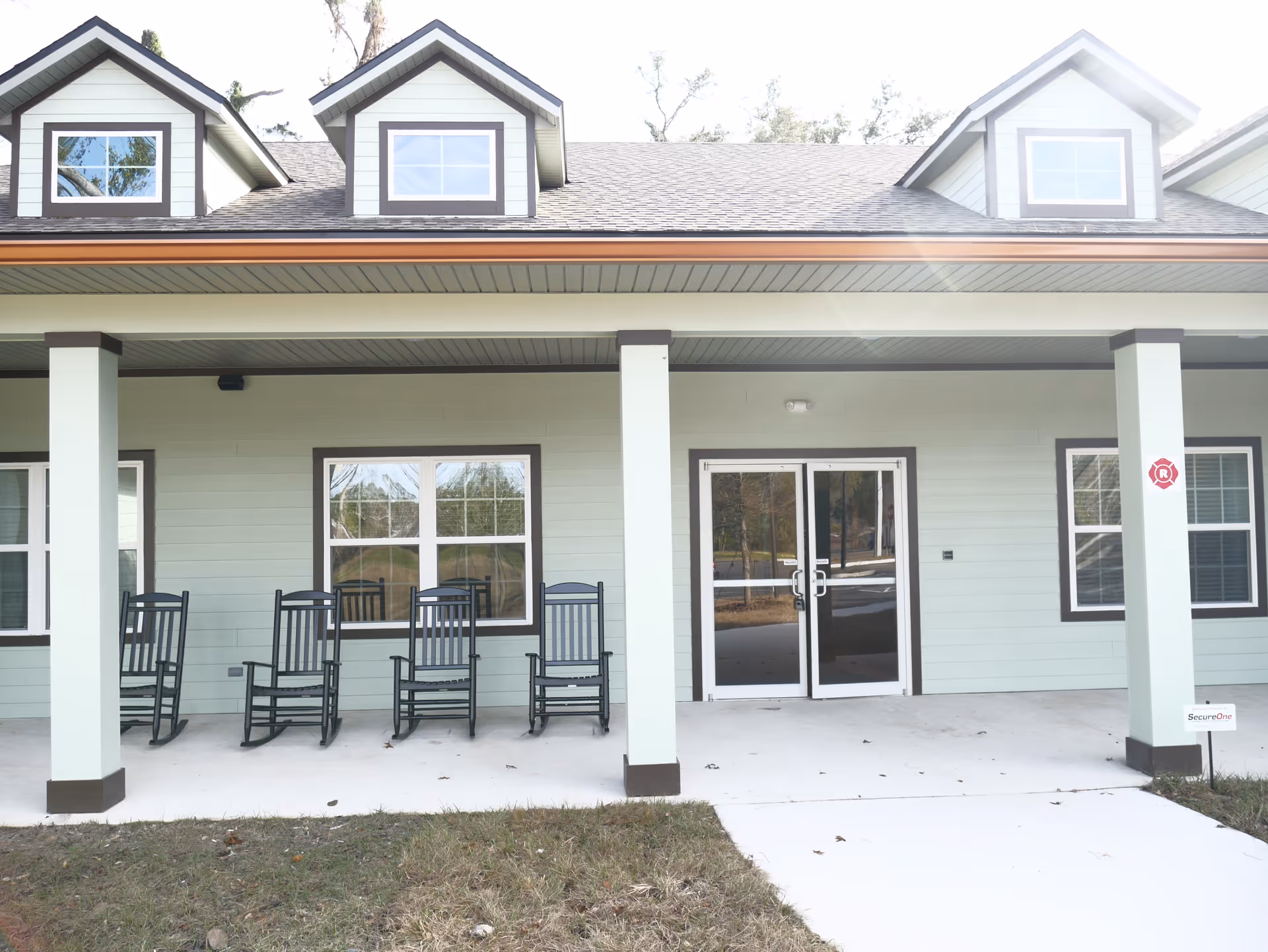 Front porch of a light-green building with several rocking chairs and glass double doors under a covered entry.