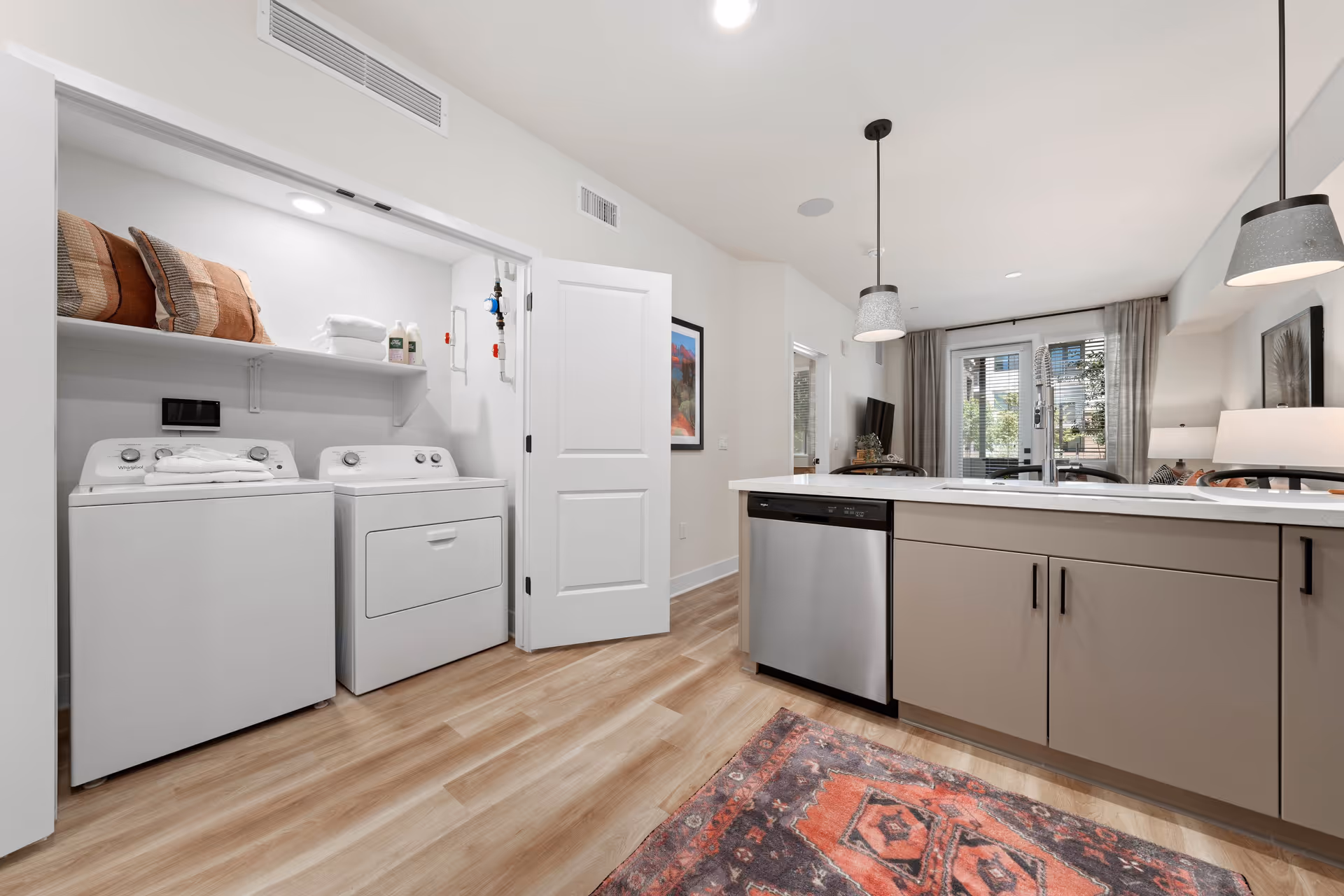 Modern kitchen area with a dishwasher, sink, and pendant lights above the counter. To the left, there is a laundry closet with a washing machine and dryer, shelves with folded towels and pillows. The room has light wood flooring and a colorful rug in front of the kitchen island. In the background, there is a living area with a TV and large windows with curtains.