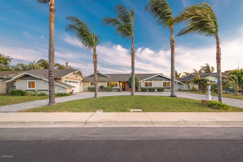 Front exterior view of a single-story residential building with a driveway, green lawn, and several tall palm trees under a partly cloudy sky.