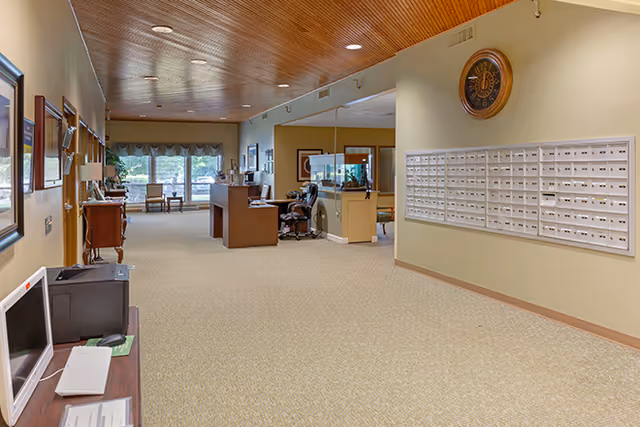 Interior view of a senior living facility lobby or common area with a reception desk, mailboxes on the wall, a clock, and seating near large windows with curtains.