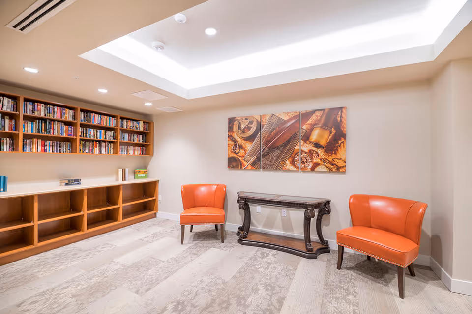 A cozy reading nook in a senior living facility featuring two orange leather chairs, a dark wooden console table, a wall-mounted triptych artwork with vintage items, and a built-in wooden bookshelf filled with books.