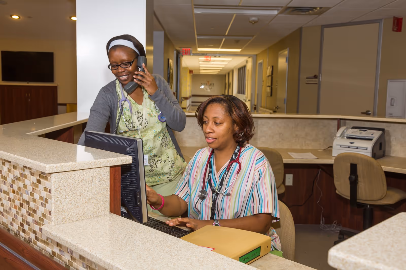 Two healthcare workers at a reception desk in a medical facility. One woman is sitting and working on a computer, wearing a striped scrub top and a stethoscope around her neck. The other woman is standing, talking on a phone, and smiling, wearing glasses, a headband, and a floral scrub top with a gray cardigan. The background shows a hallway with doors and ceiling lights.