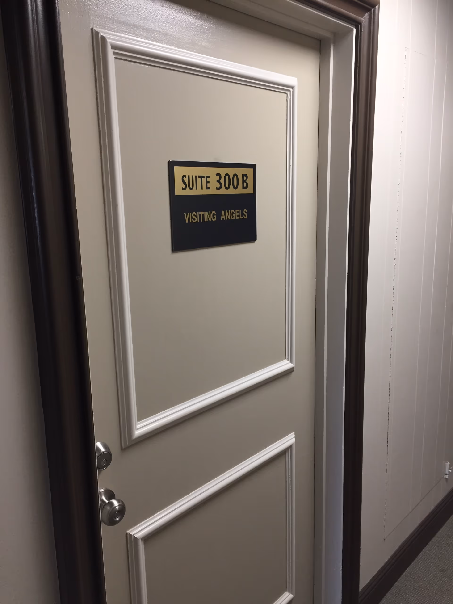 Closed beige door with white trim labeled SUITE 300 B VISITING ANGELS in a hallway with beige walls and brown trim.