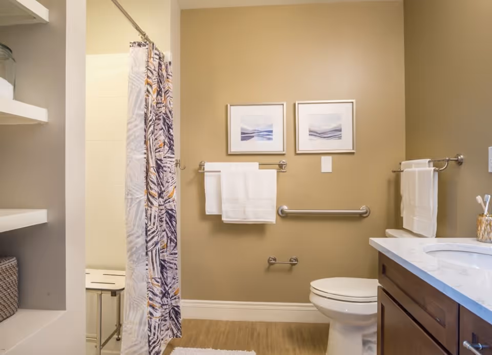 A bathroom with beige walls featuring two framed abstract art pieces above a towel rack holding white towels. There is a white toilet with a grab bar next to it, a countertop with a sink and toothbrush holder, and a shower area with a patterned curtain and a shower bench. Shelves with storage baskets are visible on the left side.