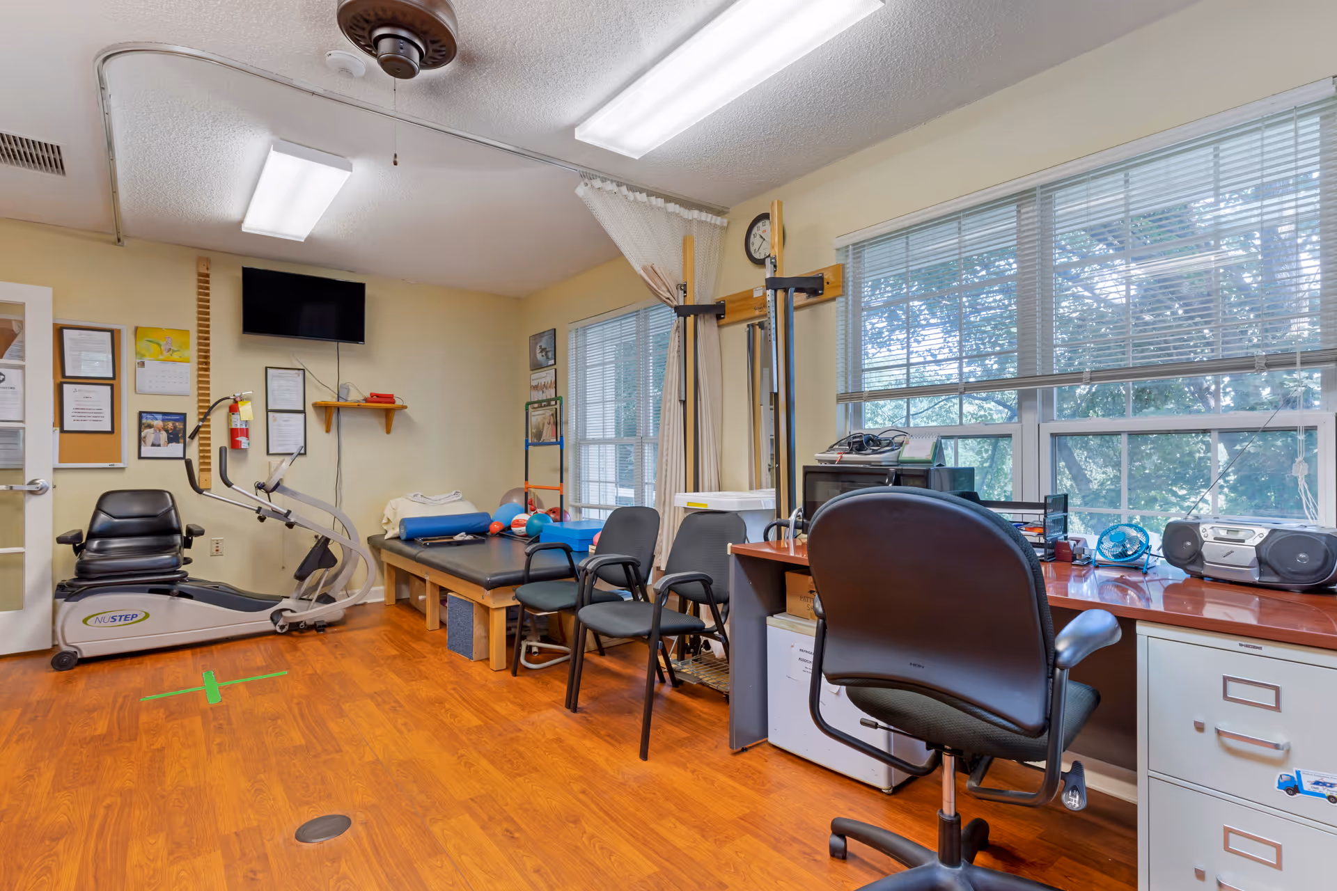 A room with exercise equipment including a NuStep machine, therapy tables with exercise balls, chairs, a desk with office supplies, and large windows letting in natural light. The room has wood flooring and light-colored walls with certificates and a TV mounted on one wall.