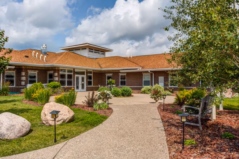Exterior view of a single-story brick building with a tan shingled roof under a partly cloudy sky. A concrete pathway leads to the entrance, surrounded by landscaped greenery, bushes, flowers, large rocks, and small garden lights. There is a wooden bench on the right side near a tree.