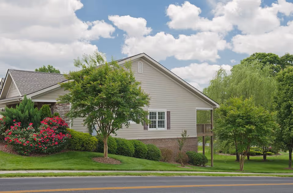 Exterior view of a single-story building with beige siding and a brick foundation, surrounded by green grass, trees, and flowering bushes under a partly cloudy sky.