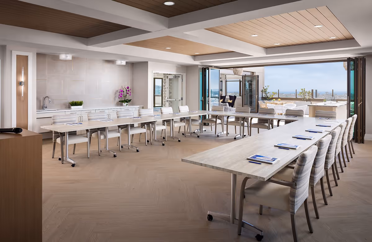 Bright meeting/dining room with U-shaped tables and chairs opening onto an outdoor terrace with ocean views.