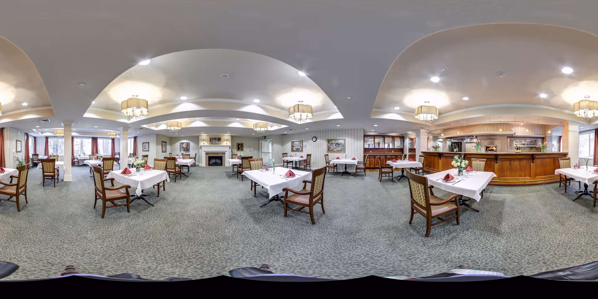 A spacious dining room with multiple tables covered in white tablecloths and set with napkins and floral centerpieces. The room features a carpeted floor, elegant ceiling lights, a fireplace, and large windows with curtains allowing natural light to enter. There is a wooden counter area on one side of the room.