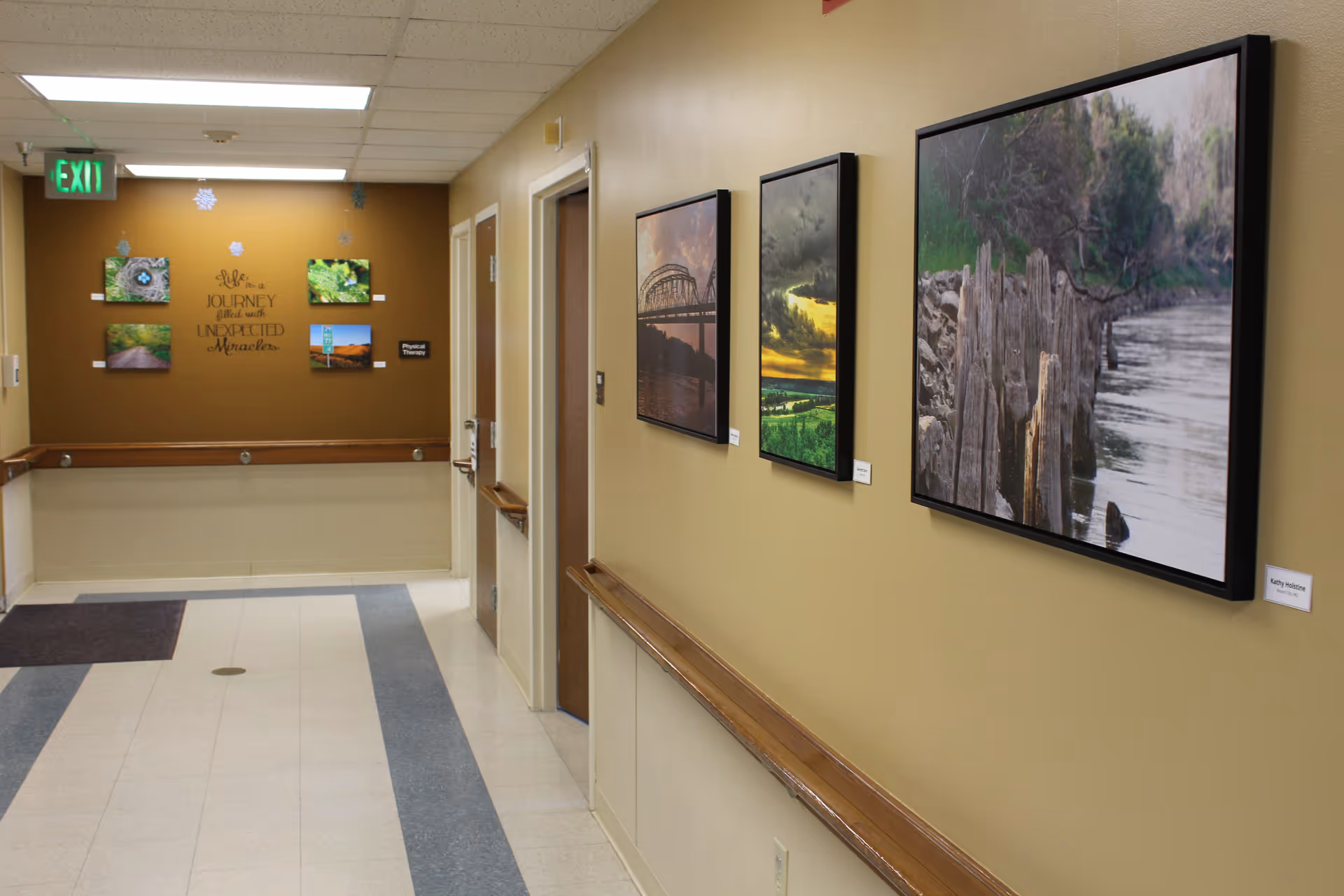 A hallway in a senior living facility with framed landscape photographs hanging on the walls. The walls are painted beige and brown, and there are handrails along the sides. At the end of the hallway, there is a sign that reads 'Physical Therapy' and a wall decal with the phrase 'Life is a journey filled with unexpected miracles.' The floor has a tiled pattern with blue and white tiles, and an exit sign is visible on the ceiling.