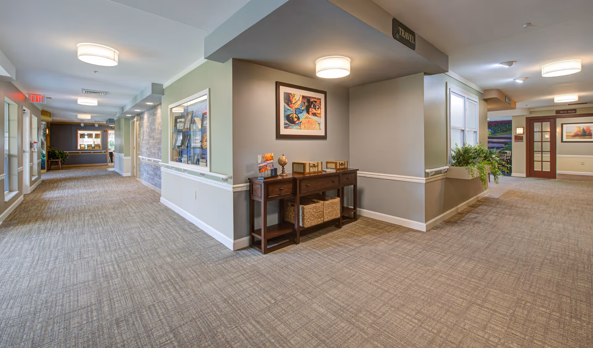 A clean, well-lit hallway in a senior living facility with beige carpet and light gray walls. The hallway features a wooden console table with decorative items and baskets underneath. There are framed artworks on the walls and potted plants near the windows. Ceiling lights illuminate the space, and exit signs are visible at the end of the corridor.