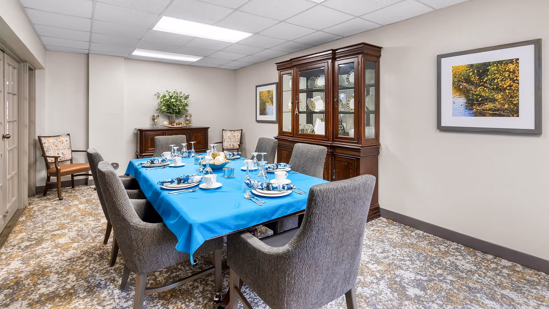 A dining room with a rectangular table covered with a bright blue tablecloth set for six people with plates, cups, silverware, and napkins. The room has six gray upholstered chairs around the table, a wooden china cabinet displaying plates, a sideboard with a floral arrangement, and framed artwork on the walls. The floor is carpeted with a patterned design.