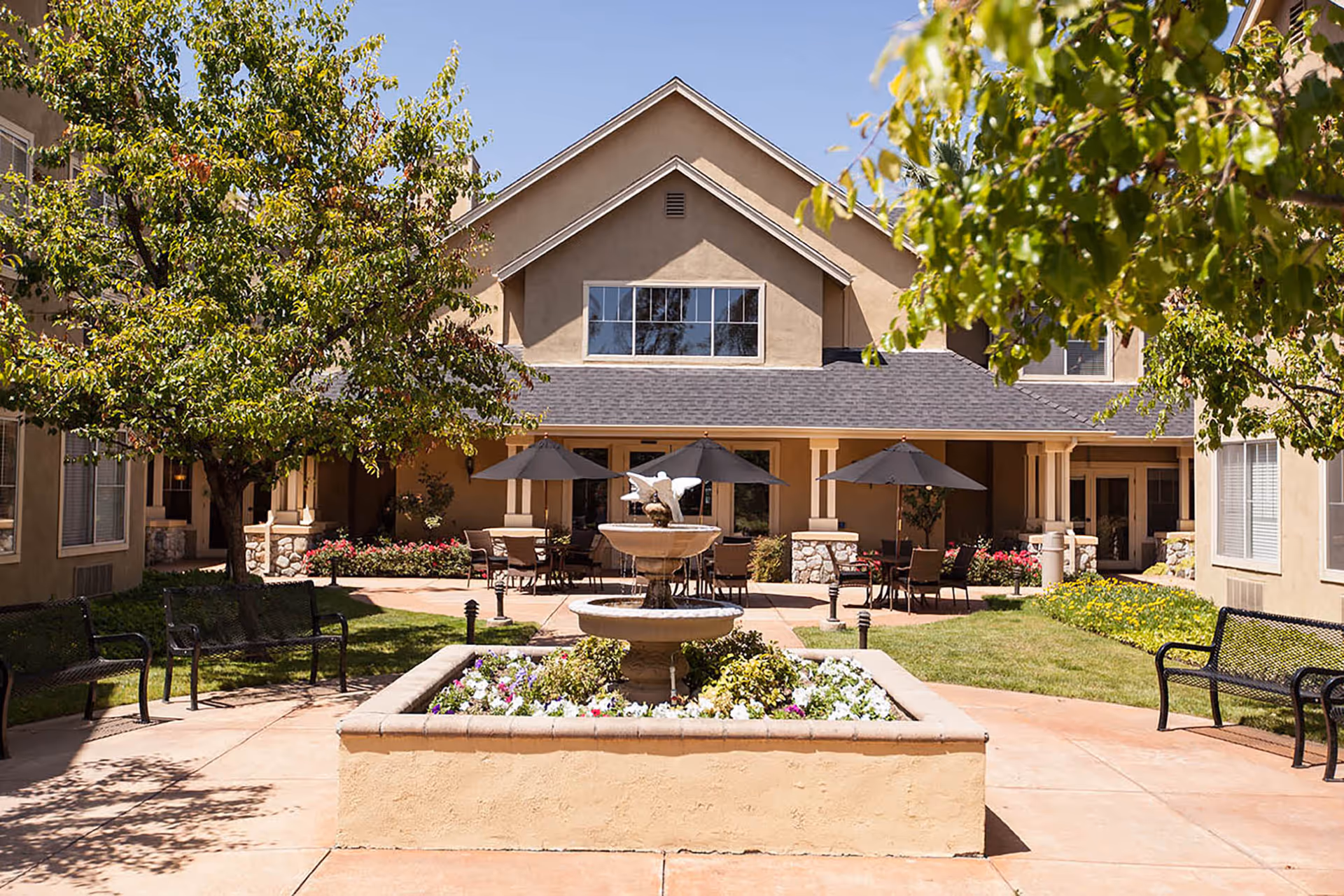 Sunny courtyard with a central fountain, flowerbeds, benches, and patio tables with umbrellas in front of a residential building.