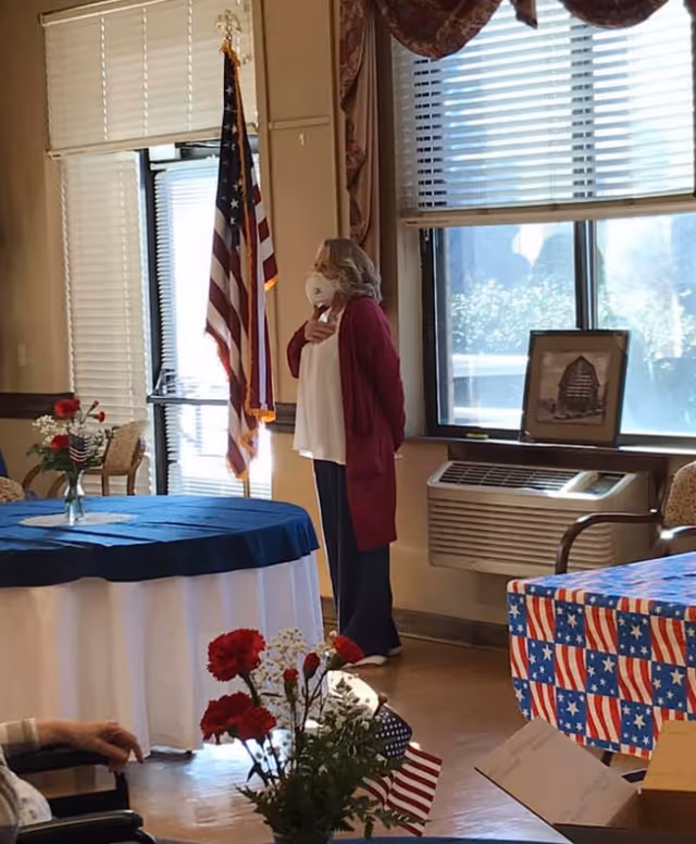 A woman wearing a face mask and a red cardigan stands with her hand over her heart in a room decorated with American flags and patriotic tablecloths. There are tables with blue and red-white-blue coverings, flowers in vases, and an American flag on a stand near a window with blinds.