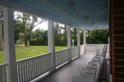 A covered veranda with white railings and rocking chairs overlooking a green lawn.