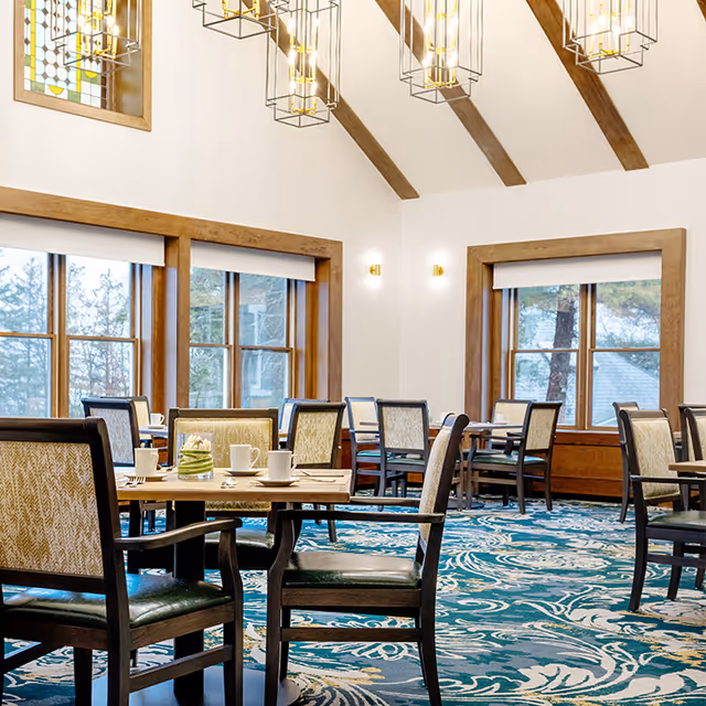 Bright communal dining room with wooden tables and chairs, large windows, vaulted ceiling with chandeliers, and patterned carpet.