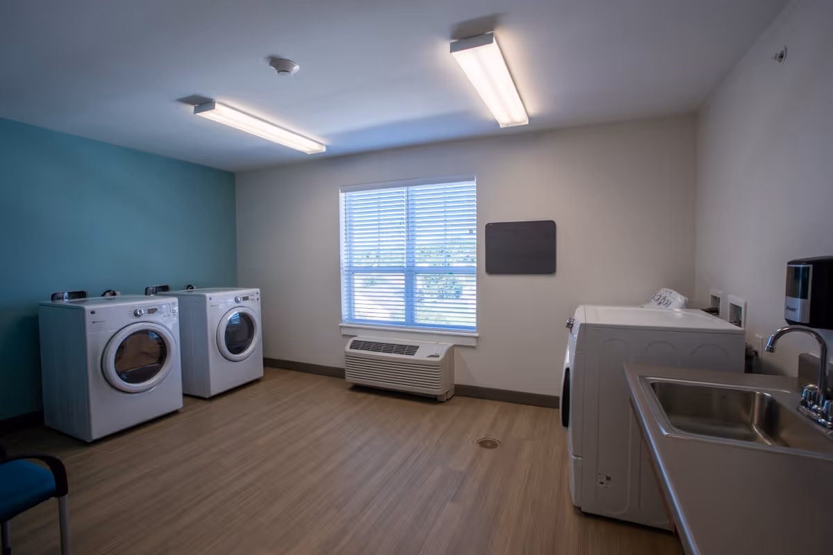 Bright communal laundry room with front-loading washers and dryers, a utility sink, window, and seating.