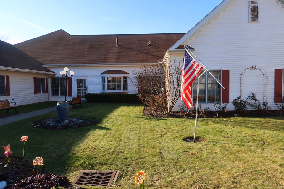 Outdoor courtyard area of a senior living facility with green grass, a flagpole displaying an American flag, benches along the walkway, a large blue vase, and a white building with red shutters in the background.