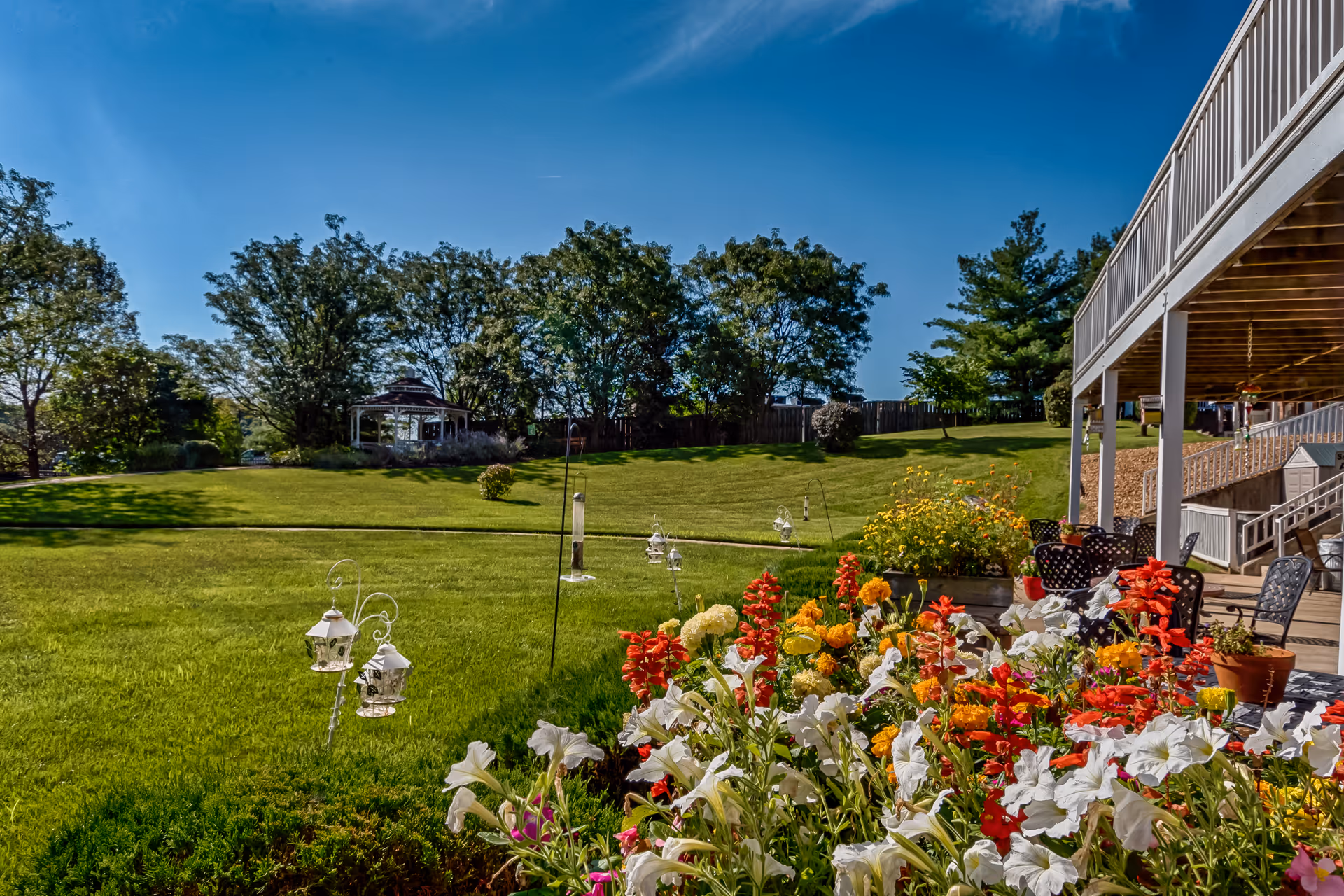 A vibrant outdoor garden area at Autumn View Gardens Assisted Living and Memory Care featuring colorful flowers in the foreground, a well-maintained green lawn, a gazebo surrounded by trees in the background, and a covered patio with chairs on the right side under a clear blue sky.
