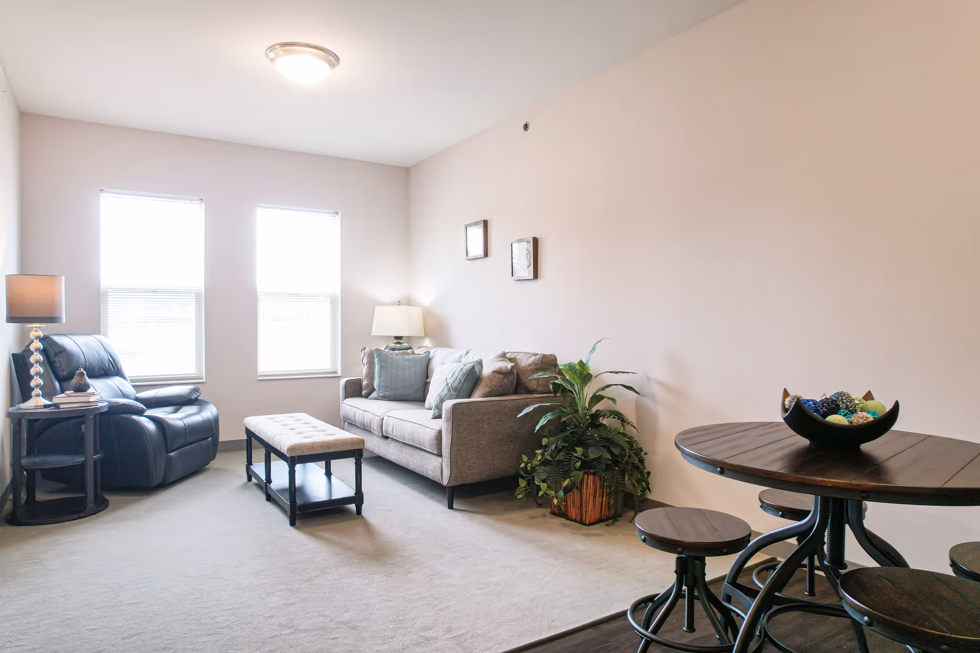 A bright living room with two large windows, a gray sofa with cushions, a black leather recliner, a small bench-style coffee table, a side table with a lamp, a potted plant, and a round wooden dining table with three stools and a decorative bowl on top.