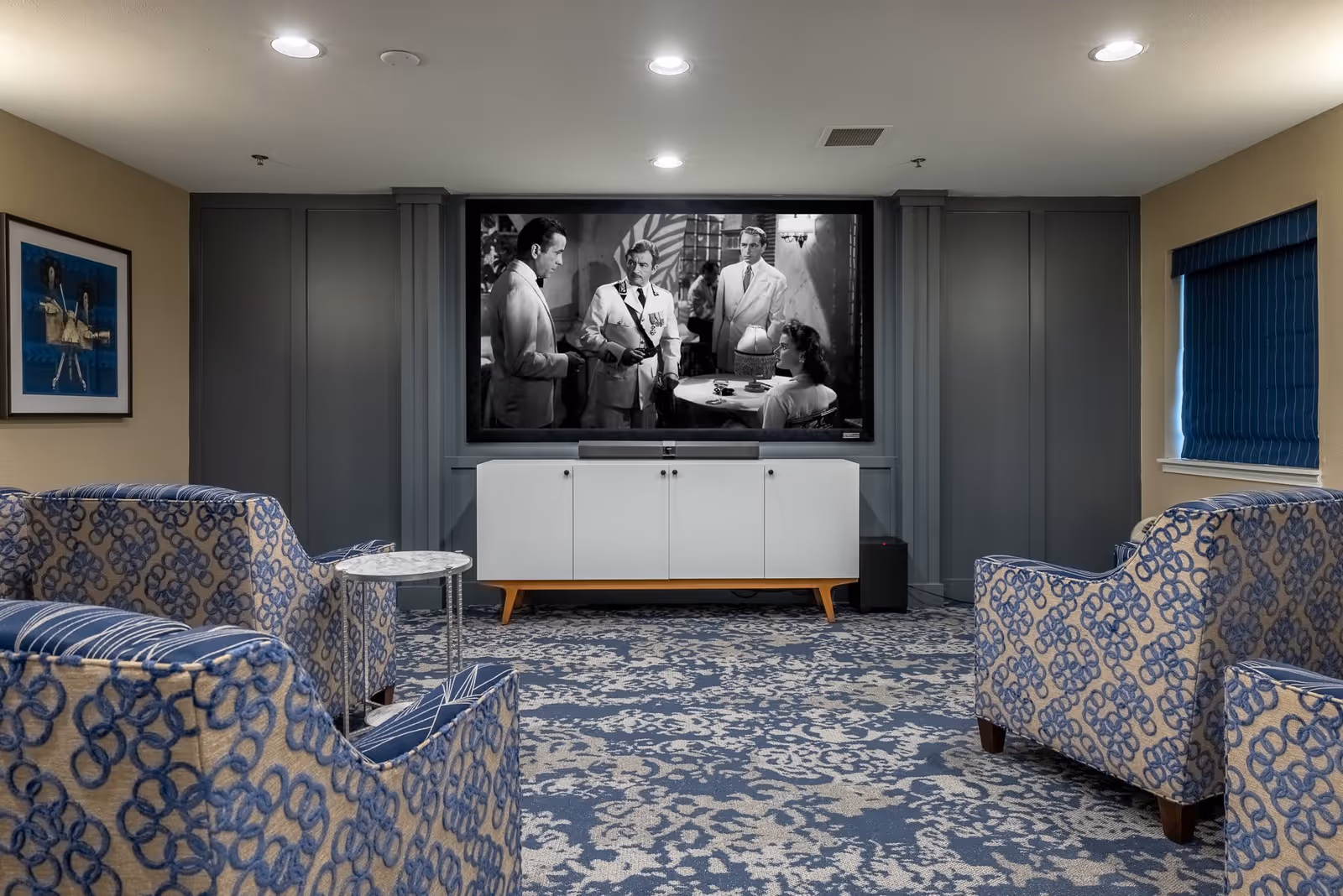 A cozy living room with patterned blue and beige armchairs arranged facing a large flat-screen TV mounted on a gray paneled wall. Below the TV is a white cabinet with wooden legs. The room has a blue and beige patterned carpet, recessed ceiling lights, a small round side table, a framed artwork on the left wall, and a window with blue blinds on the right.