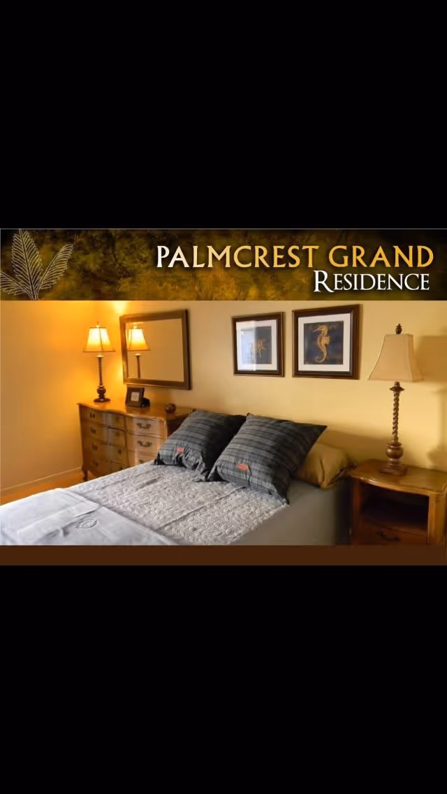 A cozy bedroom in Palmcrest Grand Residence featuring a neatly made bed with two plaid pillows, a wooden dresser with a lamp and framed mirror above it, two framed seahorse pictures on the wall, and a wooden nightstand with a lamp.