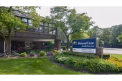 Exterior view of a health services building with a sign in front that reads ManorCare Health Services. The building is surrounded by green grass, bushes, and trees under a clear sky.