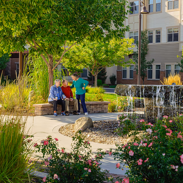 Three elderly people sitting and standing near a stone bench in a garden area with trees, flowers, and a water fountain in front of a multi-story residential building.