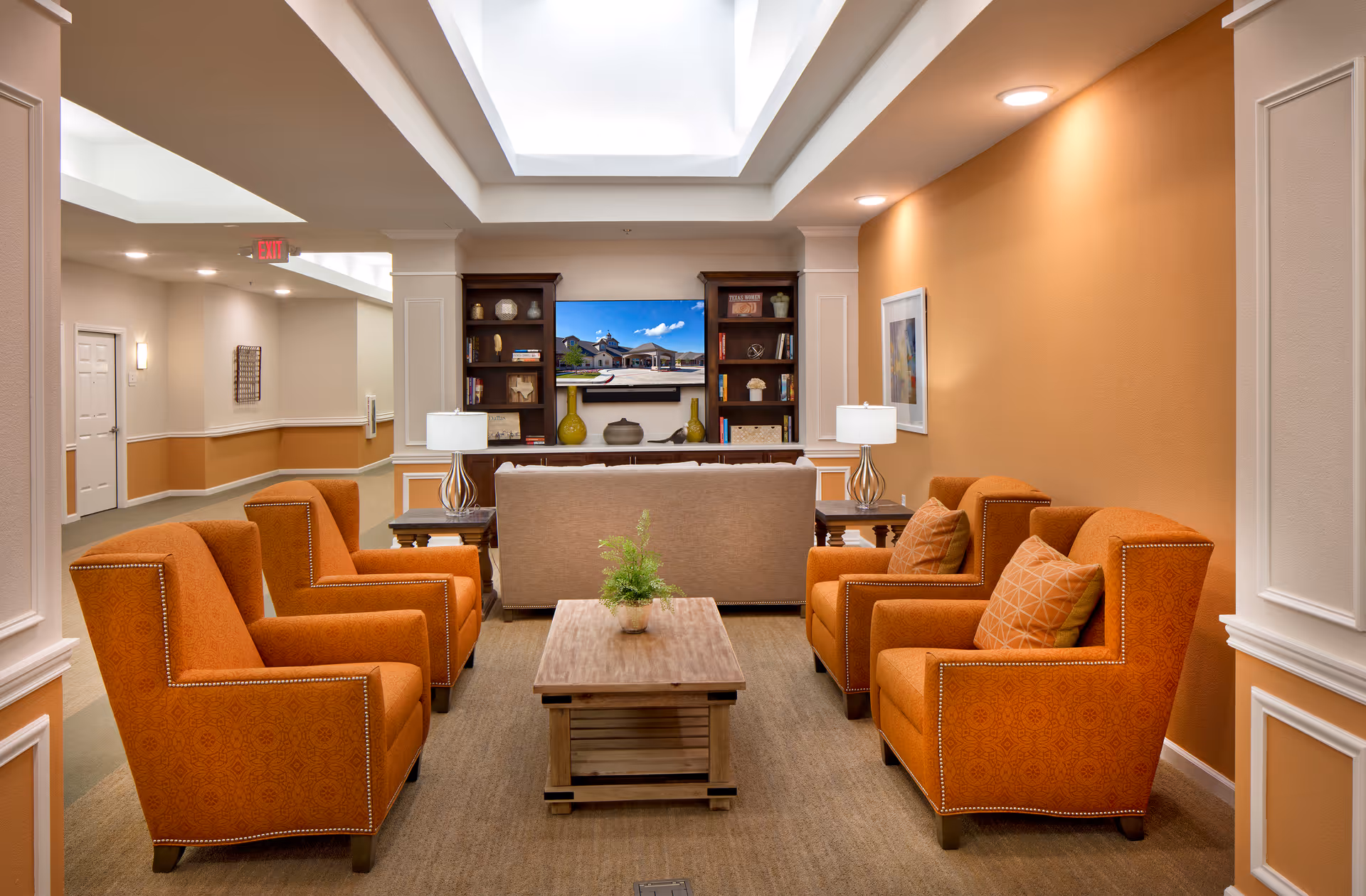 A cozy sitting area in an assisted living facility with four orange armchairs arranged around a wooden coffee table with a small plant. Behind the chairs is a beige sofa facing a TV mounted on a wall between two dark wooden bookshelves. The walls are painted in warm tones with white trim, and the ceiling has recessed lighting and a skylight.