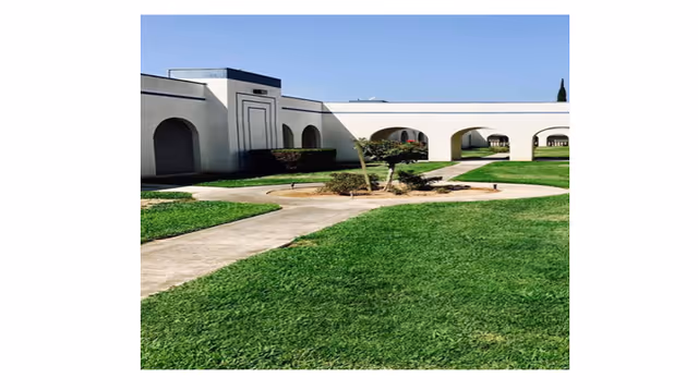 Outdoor view of a retirement center with well-maintained green lawns, concrete walkways, and a white building featuring multiple archways under a clear blue sky.