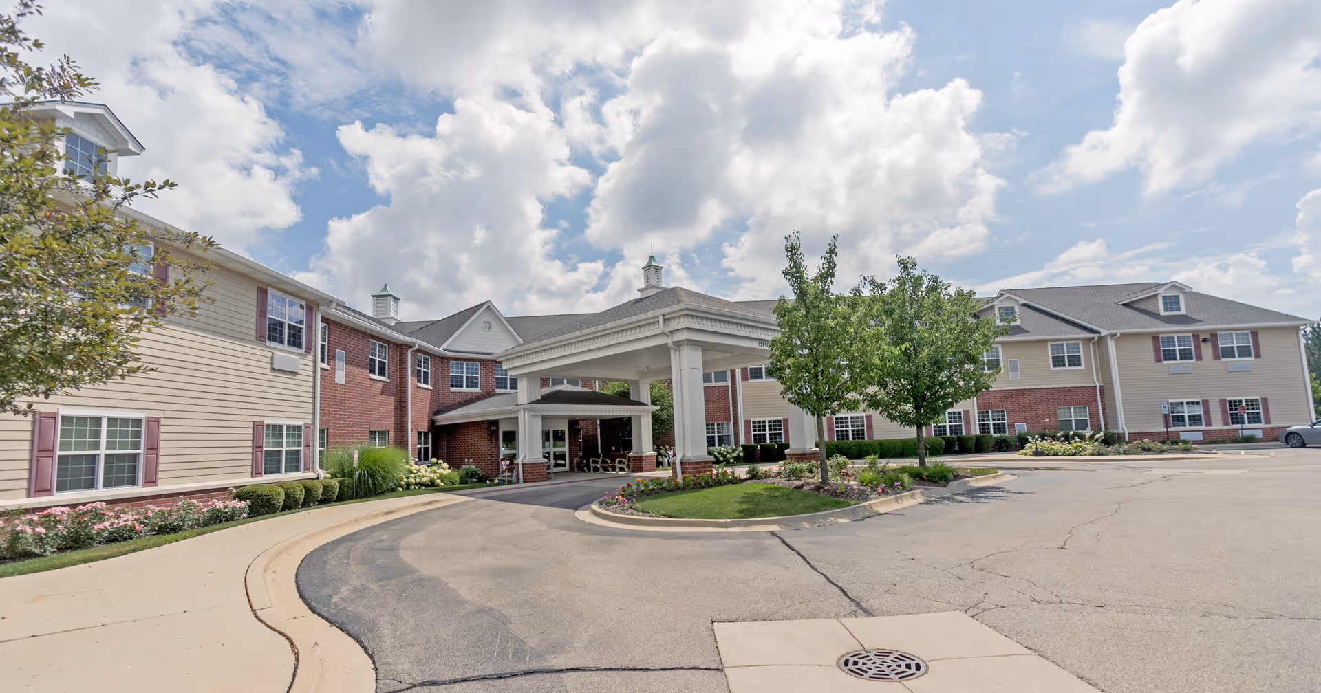 Exterior view of Heritage Woods of Huntley senior living facility showing a large building with a covered entrance, landscaped greenery, trees, and a partly cloudy sky.